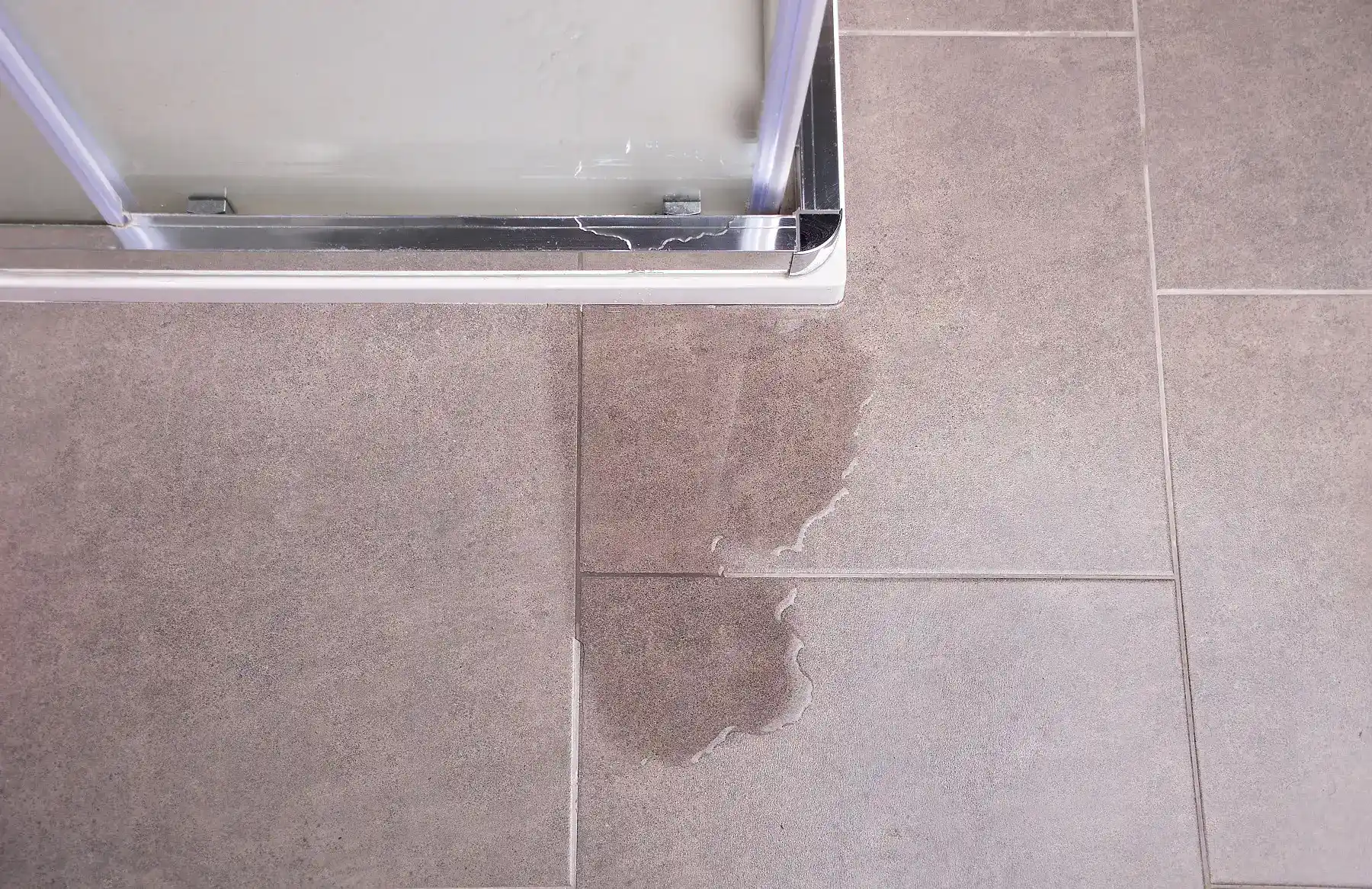 Water puddle outside framed shower door shows failed bottom seal on large gray tile floor.